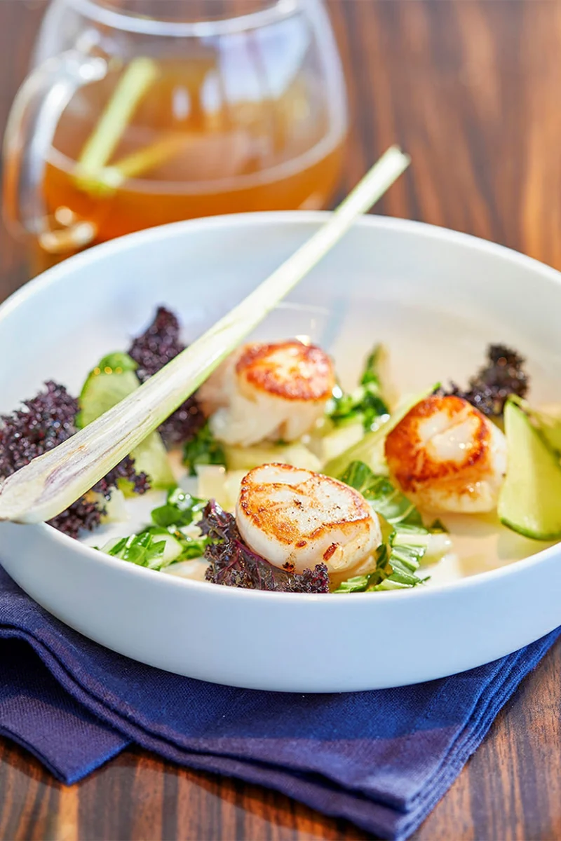 Trois noix de Saint-Jacques dorées dans une assiette creuse, chou kale violet, bok choy, lamelles vertes et citronnelle