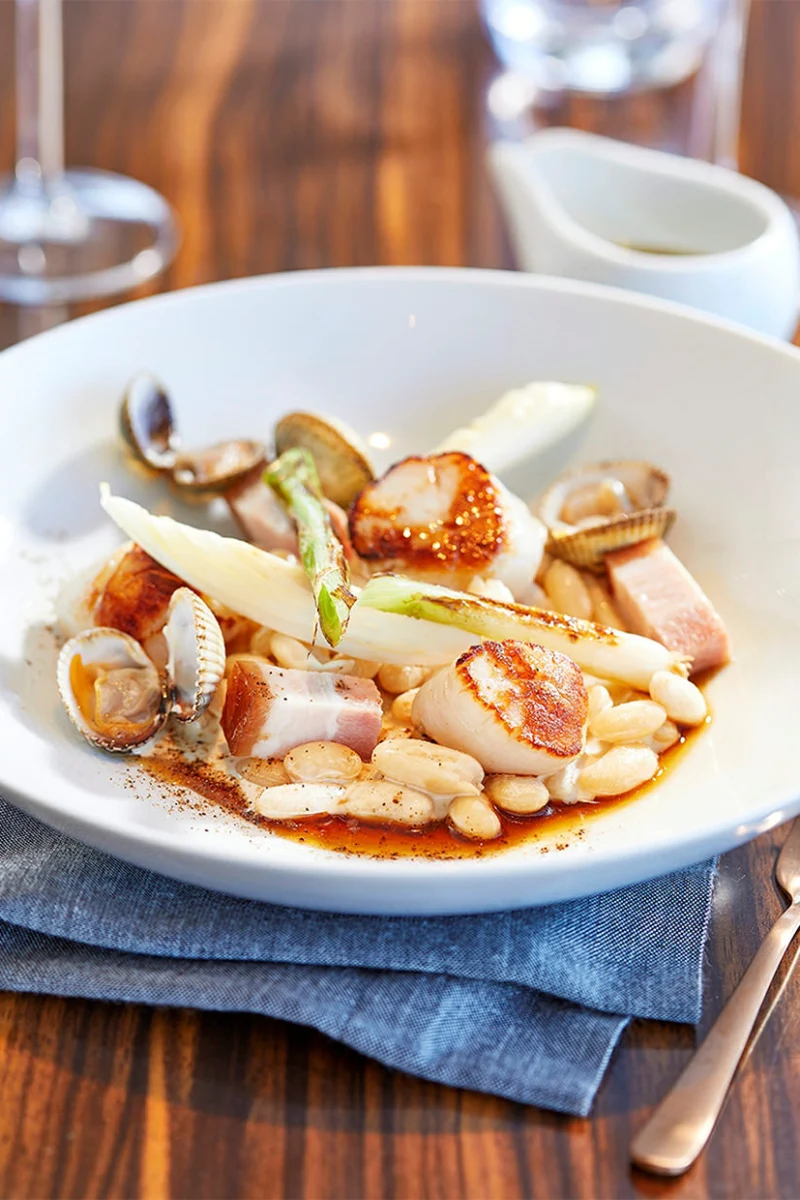 Noix de Saint-Jacques snackées sur des cocos de Paimpol, coques, lard, endive et cébette dans une assiette creuse