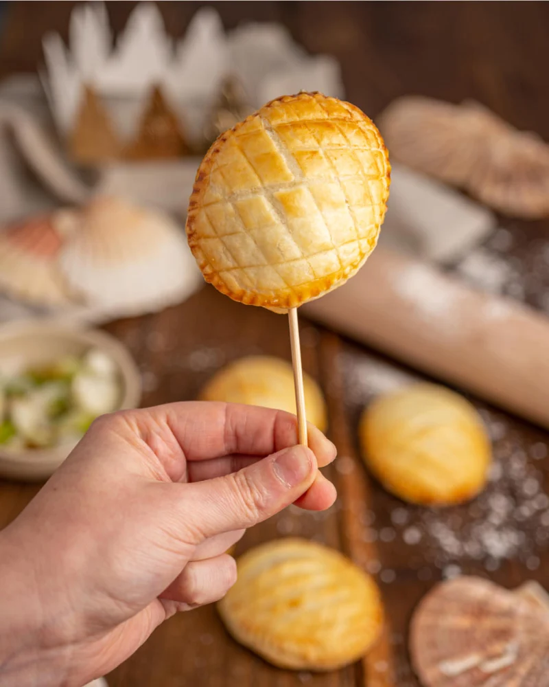 Mini-galette feuilletée dorée et quadrillée piquée sur un bâton, tenue à la main, coquilles Saint-Jacques en arrière-plan