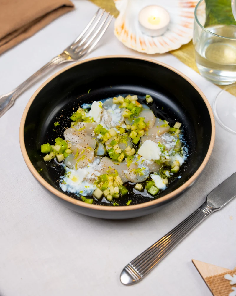 Lamelles de noix de Saint-Jacques crues, dés de céleri et de pomme verte et paillettes de granité blanc dans un bol noir