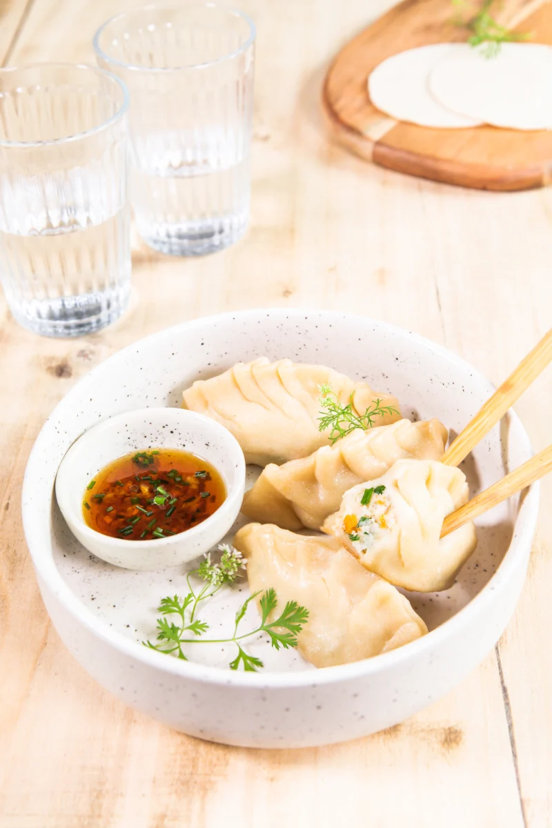 Gyozas plissés dans un bol, l’un ouvert sur sa farce, sauce soja à la ciboulette et brins de coriandre