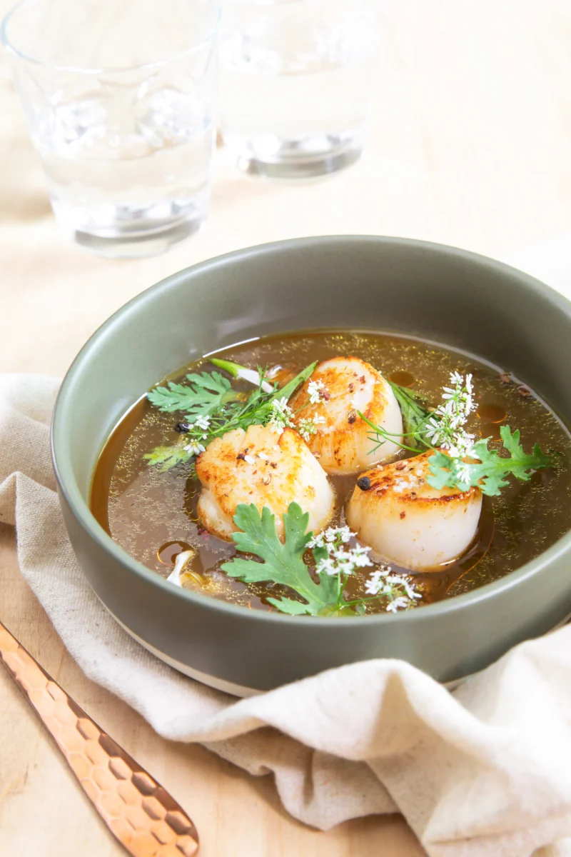 Trois noix de Saint-Jacques dorées dans un bouillon ambré, herbes fraîches et fleurs blanches, dans un bol vert kaki