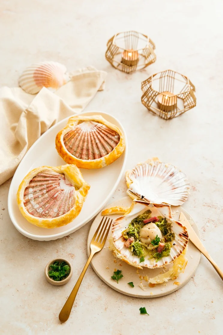 Coquilles Saint-Jacques soudées de pâte feuilletée dorée, l’une ouverte sur une noix, du chou frisé, des lardons et du persil
