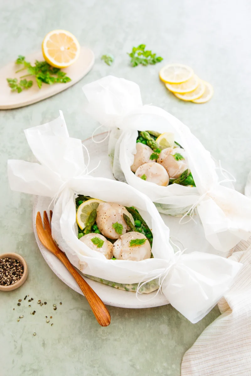 Papillotes ouvertes de noix de Saint-Jacques, asperges vertes, petits pois et rondelle de citron, feuilles de cerfeuil