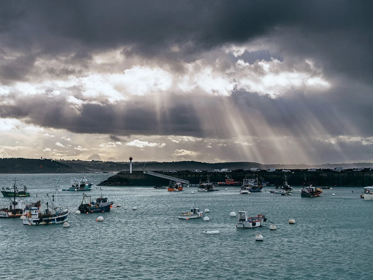 Bateaux de pêche au mouillage devant la jetée et le phare d'un port, sous un ciel d'averse percé de soleil
